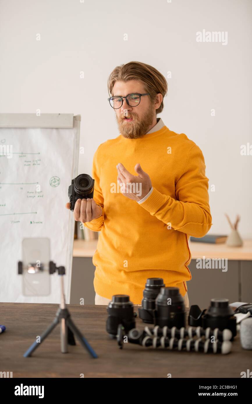 Contemporary young professional photographer holding photocamera and explaining how it works to his online audience at lesson Stock Photo