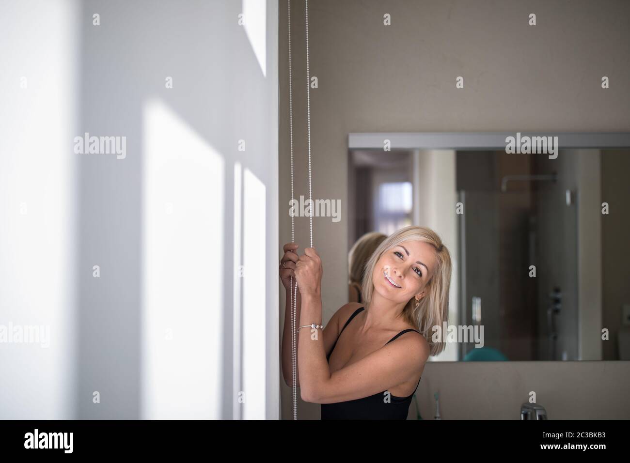 Pretty, young woman lowering the interior shades/blinds in her modern interior apartment Stock Photo