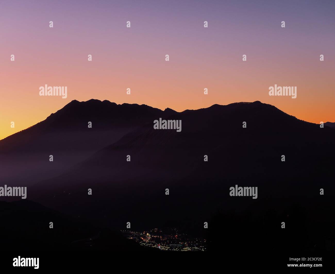 View of a town glowing in the distance surrounded by mountains against the sunset sky Stock Photo