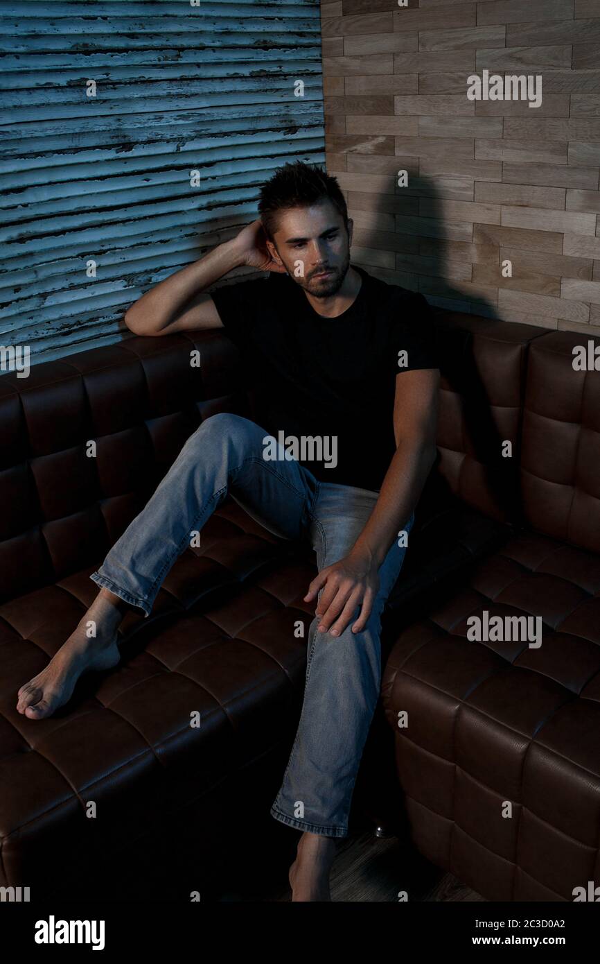 portrait of a young Caucasian guy in a black t shirt and gray jeans sitting on a sofa in a stylish interior Stock Photo
