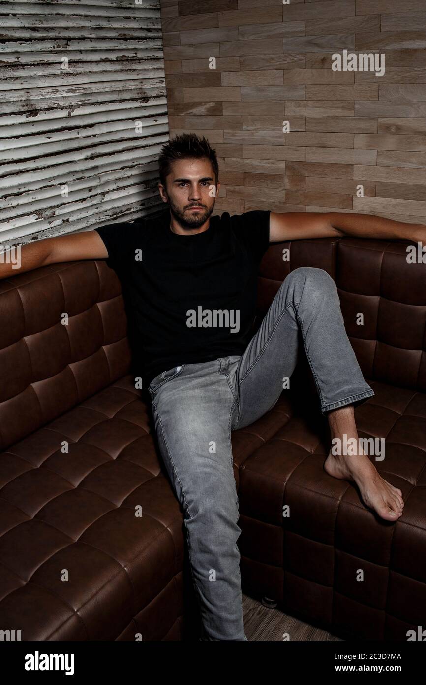 portrait of a young Caucasian guy in a black t shirt and gray jeans sitting on a sofa in a stylish interior Stock Photo