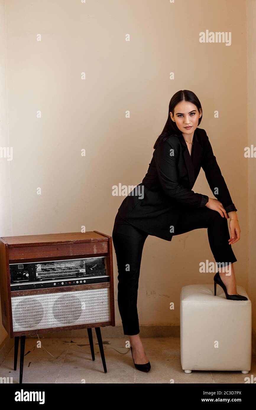 a beautiful Caucasian young woman in a black pantsuit and black sandals stands next to a vintage record player Stock Photo