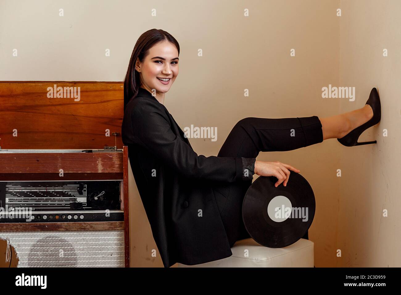 a beautiful Caucasian young woman in a black pantsuit and black sandals poses next to a vintage record player with a gramophone Stock Photo