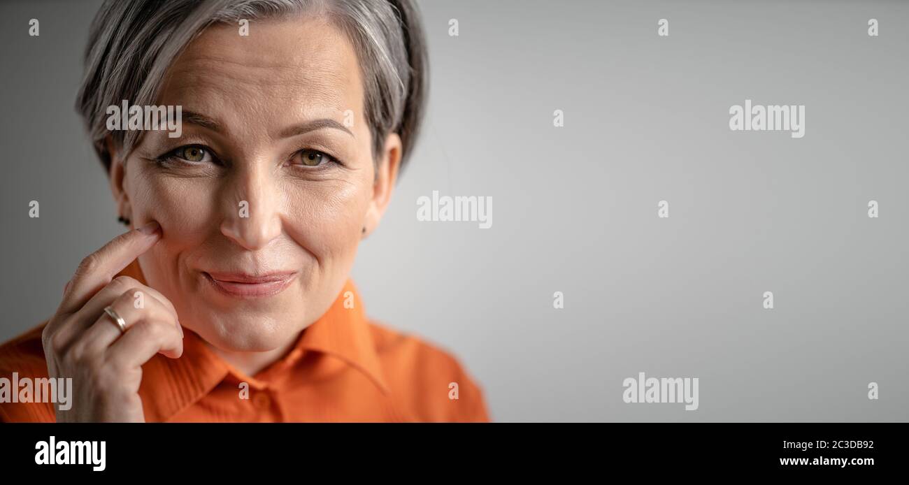 Charming gray-haired woman smiles touching cheek with finger. Selective focus on female face with tiny wrinkles. Close up portrait. Mature skin care Stock Photo