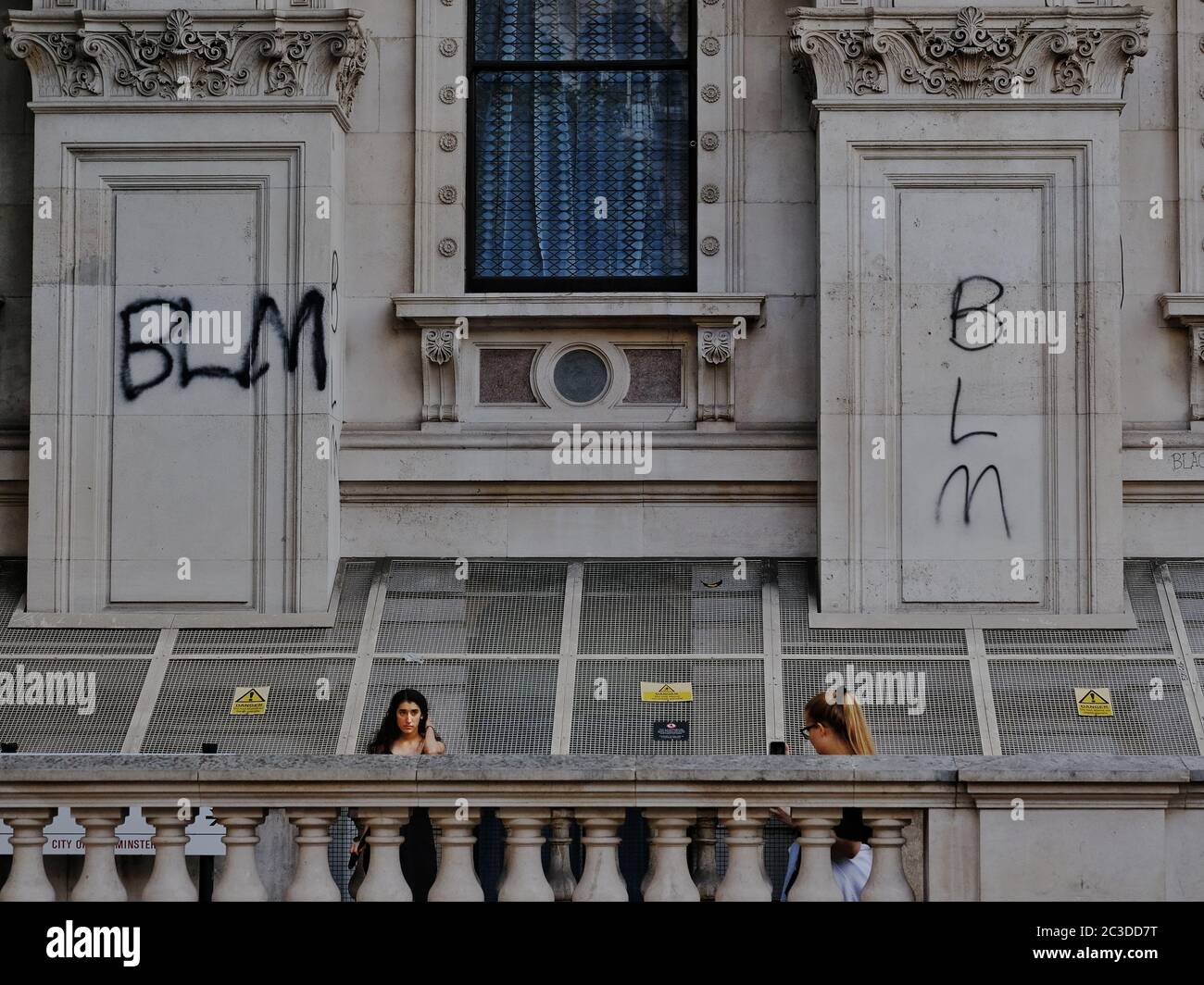 Two young women take photos of each other in Whitehall with BLM (Black Lives Matters) graffiti spraypainted on the building behind. Stock Photo