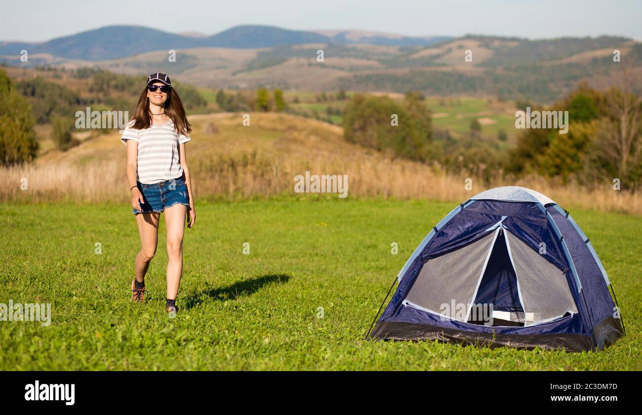 Lifestyle image of happy woman walking near tent with mountains background. Wearing stylish sunglasses, jeans shorts and shirt. Freedom, tourism conce Stock Photo