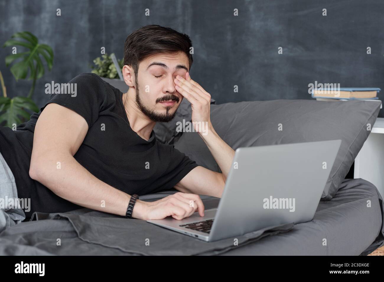 Tired young man having eye fatigue lying on bed with laptop and rubbing eye Stock Photo