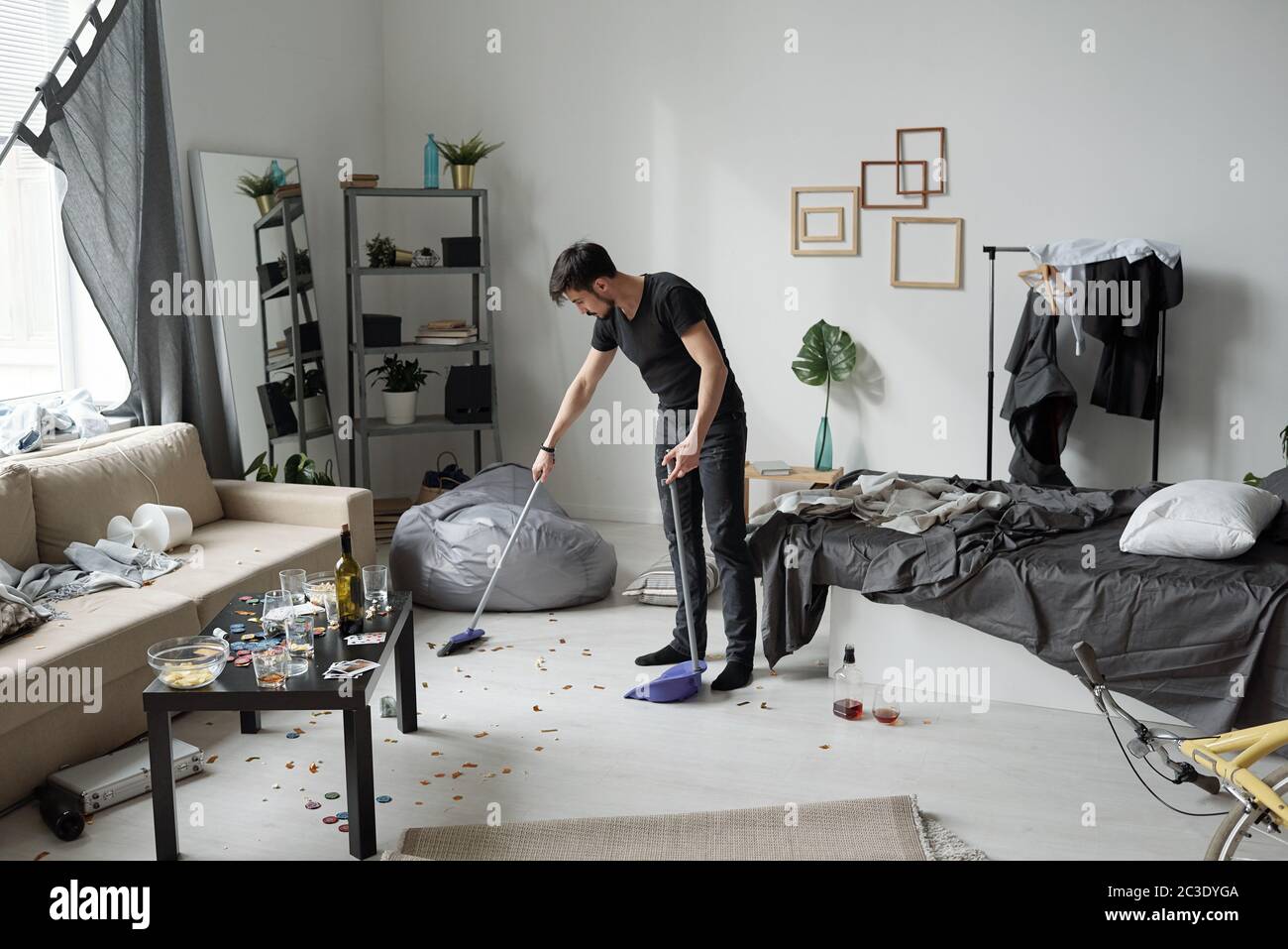 Young man sweeping floor at home after party: bottles, crumps and poker chips on floor Stock Photo