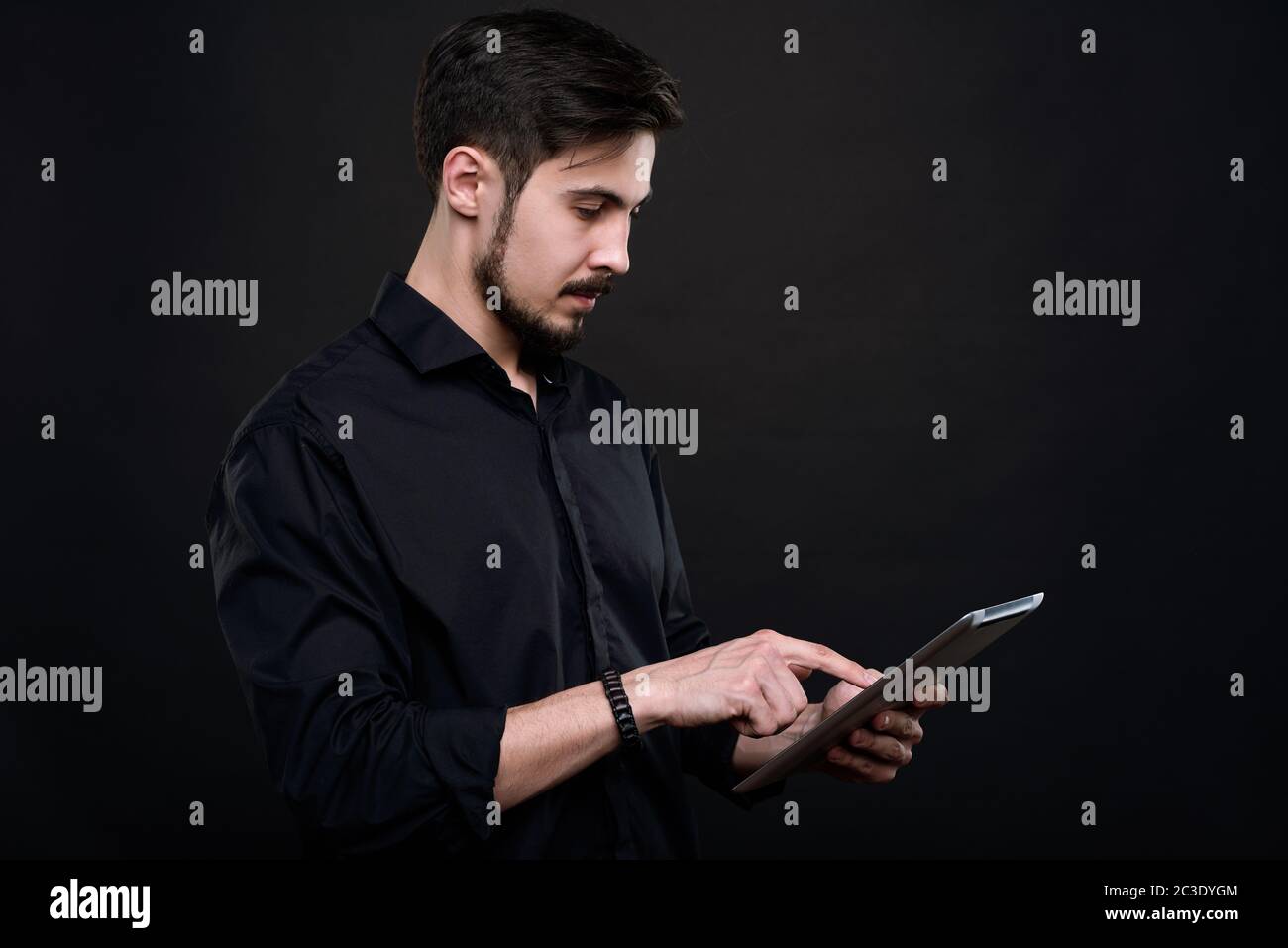 Serious young bearded man standing against black background and analyzing data on tablet Stock Photo