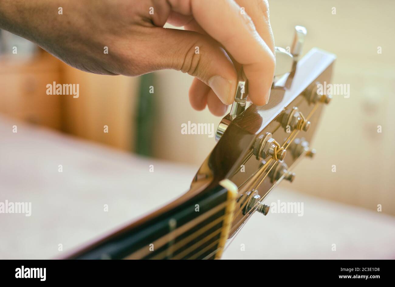 Tuning the guitar. Fingers are turning the tuning peg on the head of acoustic guitar. Authentic shot with blurred room in the background. Stock Photo