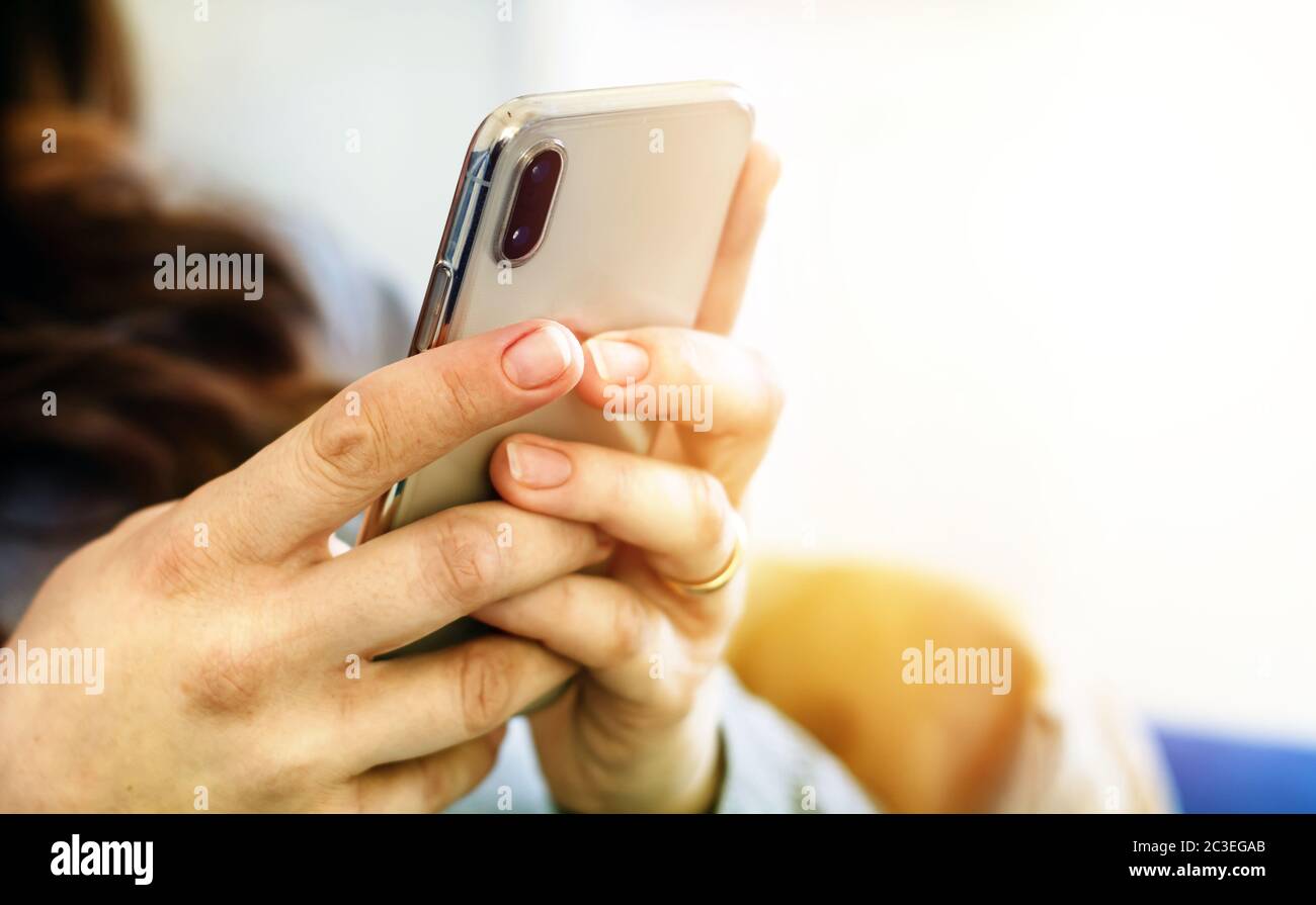 the hands of a young caucasian brunette woman while holding a smartphone typing on the touch screen. Stock Photo