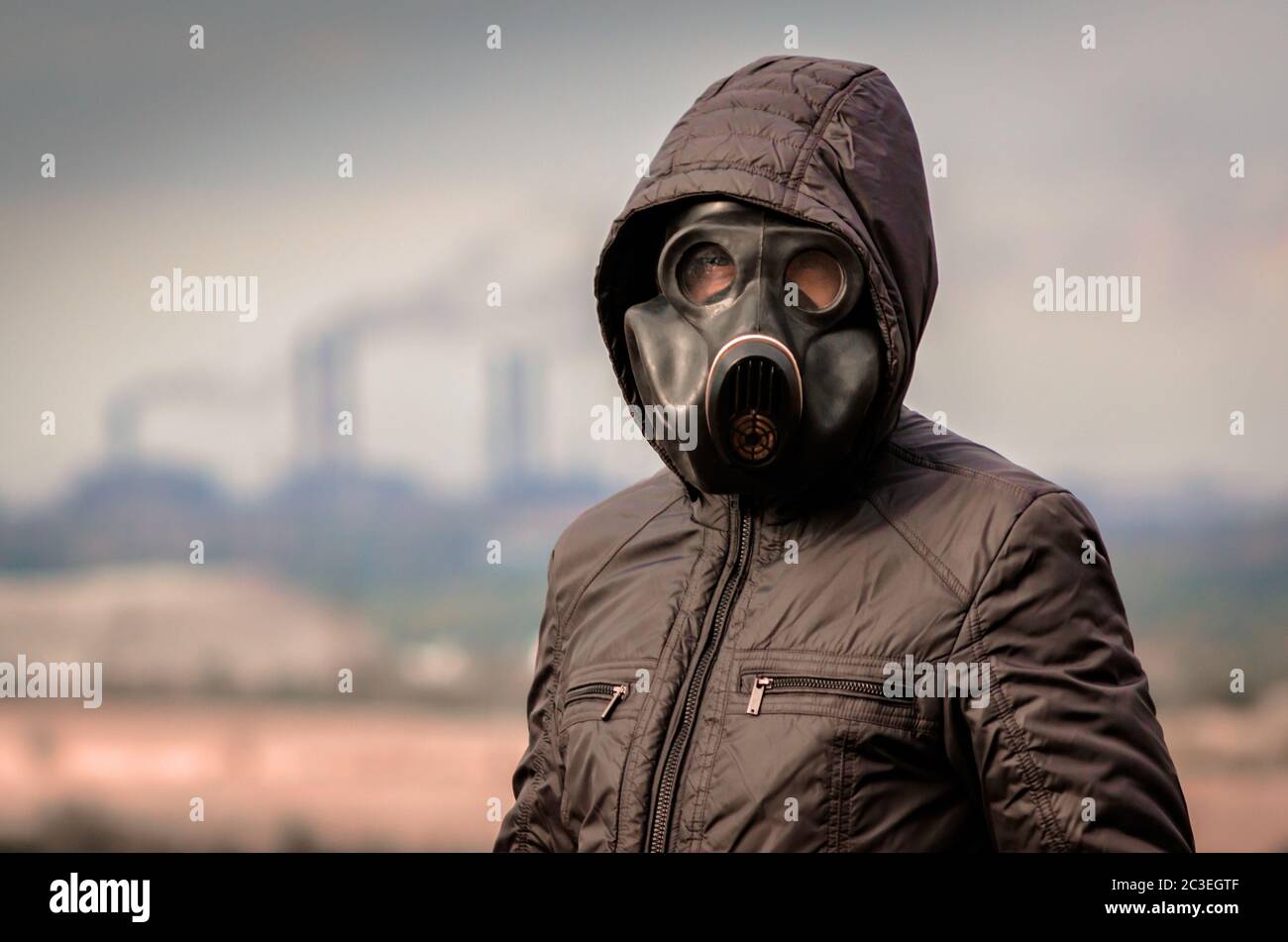 portrait of a man in a gas mask and a hood against the background of smoke from factory pipes