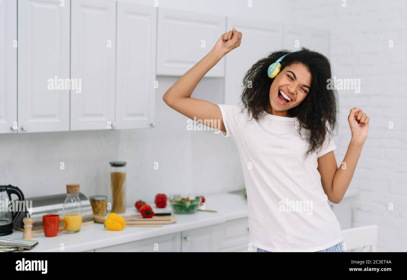 Happy emotional woman dancing at home, having fun. Portrait of beautiful African American female listening music using wireless headphones Stock Photo