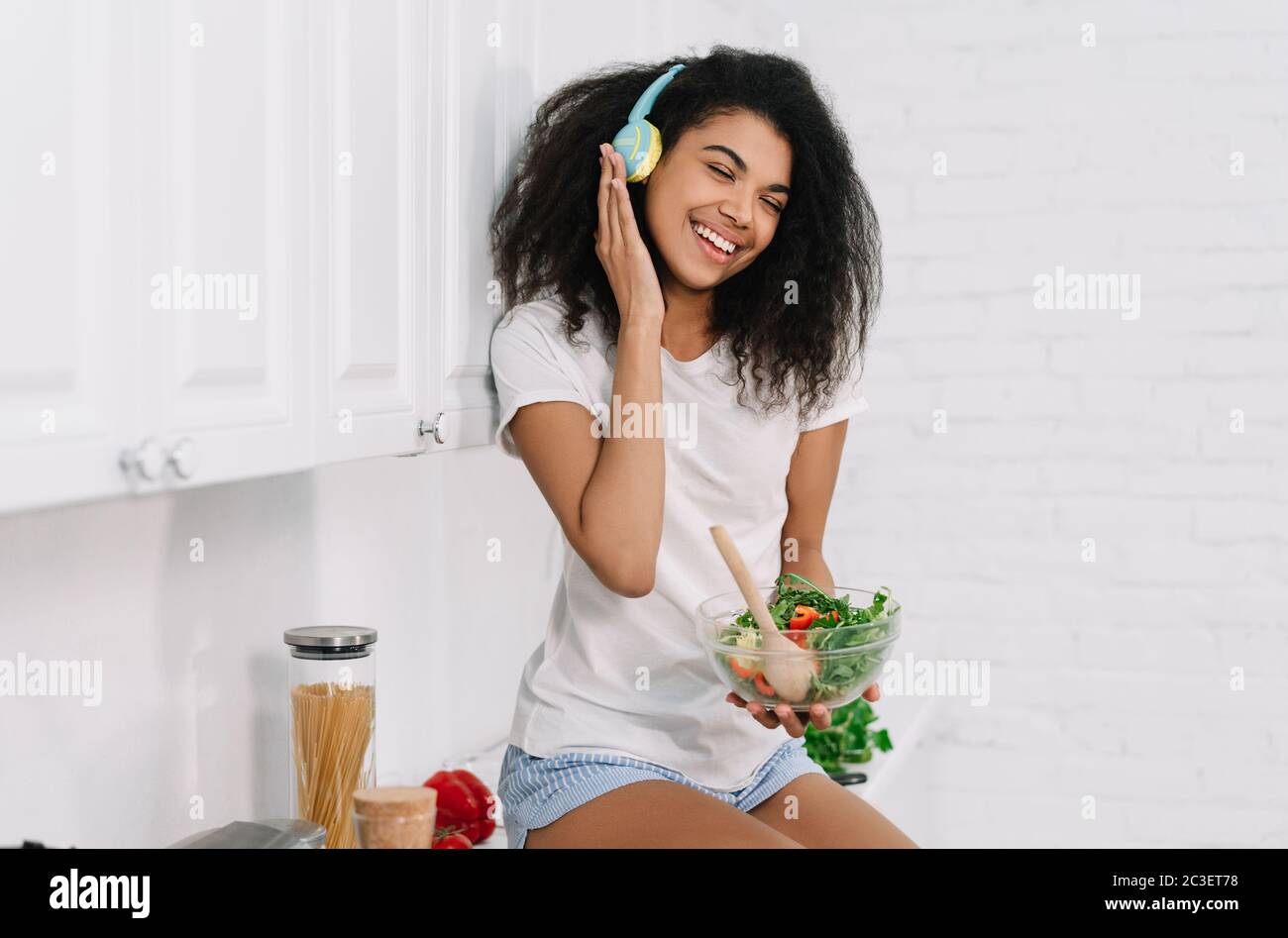 Happy emotional girl holding bawl with fresh salad, listening music at home, laughing Stock Photo