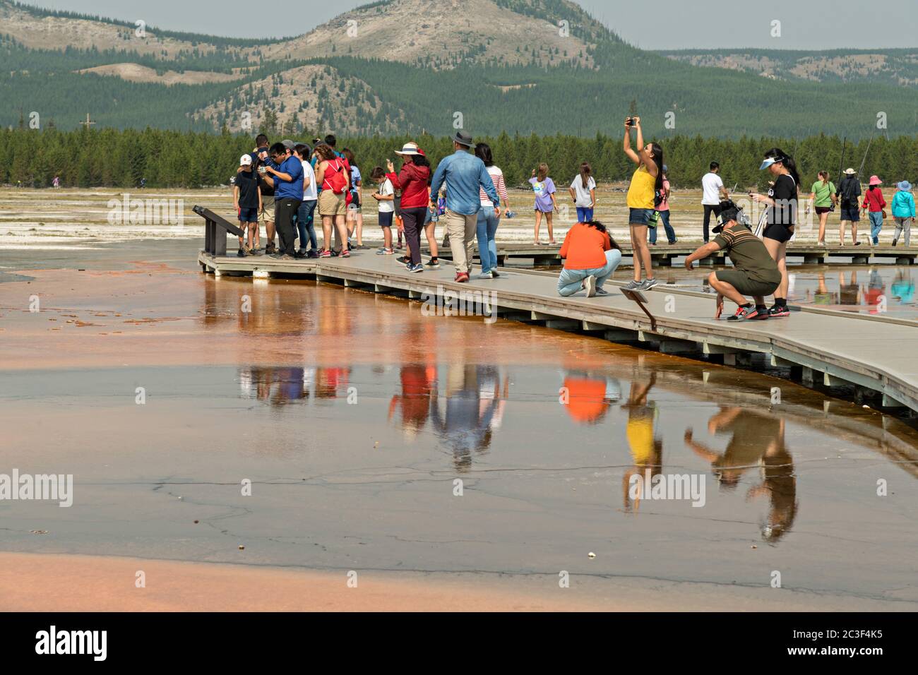 Tourists walk along the boardwalk at Grand Prismatic Spring the largest hot spring in Yellowstone National Park and third largest in the world. Grand Prismatic is about 250 by 300 feet in size, averages 160 degrees Fahrenheit and is up to 160 feet deep. The bright colors around the spring are from cyanobacteria mats. The Grand Prismatic Spring is part of the Midway Geyser Basin Excelsior Group in Yellowstone, Wyoming. Stock Photo