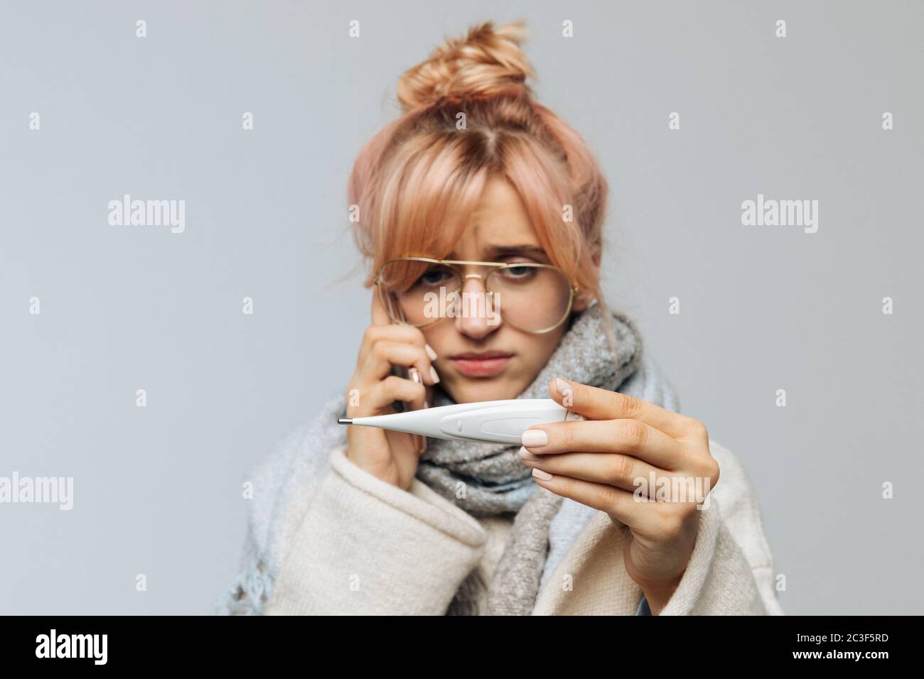 Closeup portrait of European unhealthy woman in glasses caught cold and looking at thermometer. Sick young blonde female in scarf feeling bad, talking Stock Photo