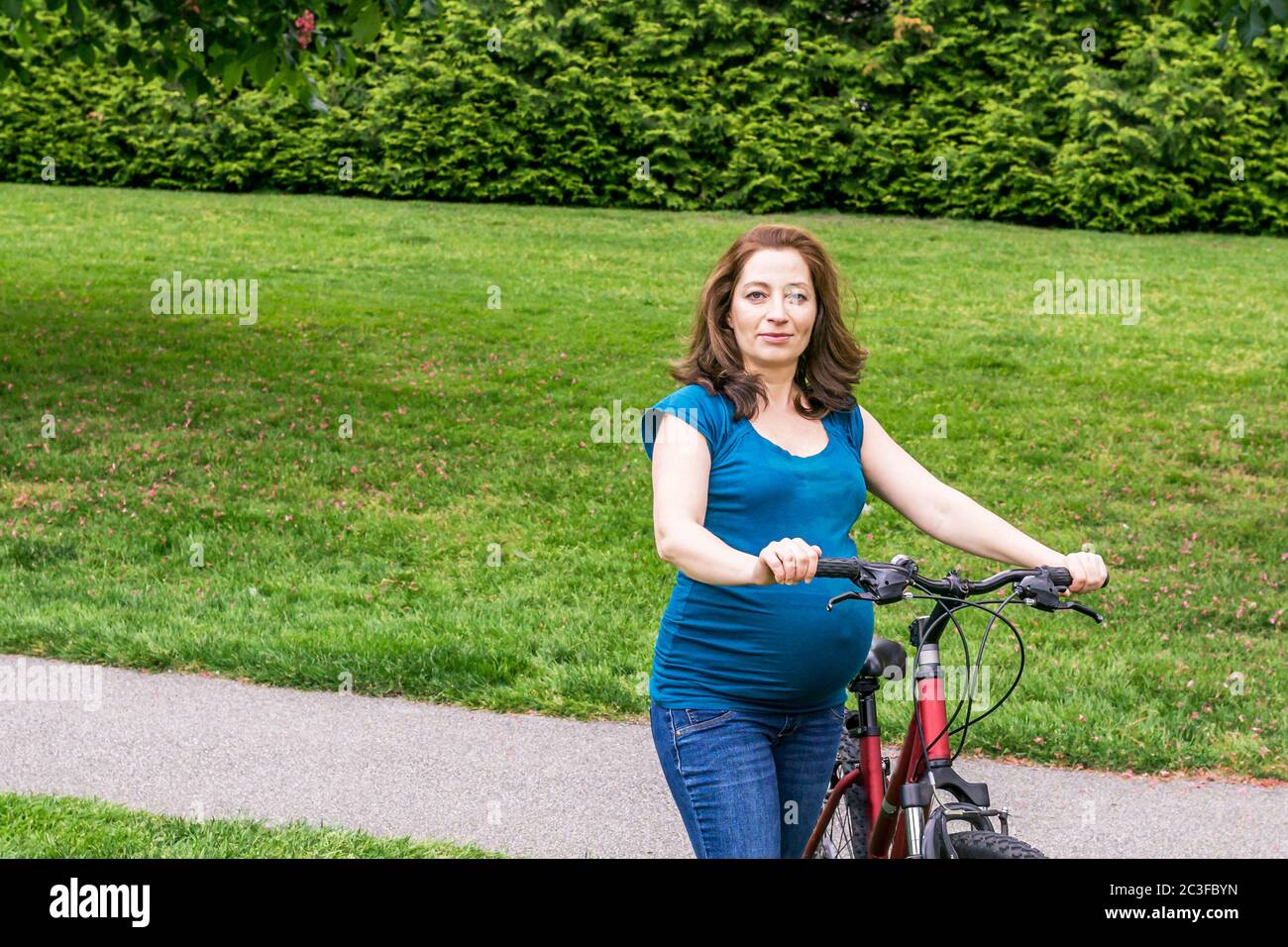 pregnant woman on the green lawn in a park holding bicycle ready to ride. Stock Photo