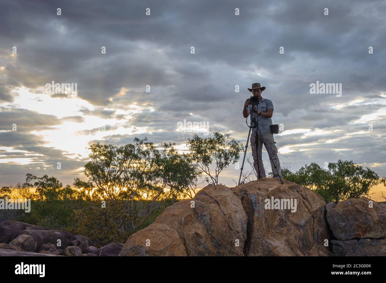 An adventure photographer sets up on a rocky ridge top to photograph a golden hour sunset landscape at Undarra in Queensland, Australia. Stock Photo