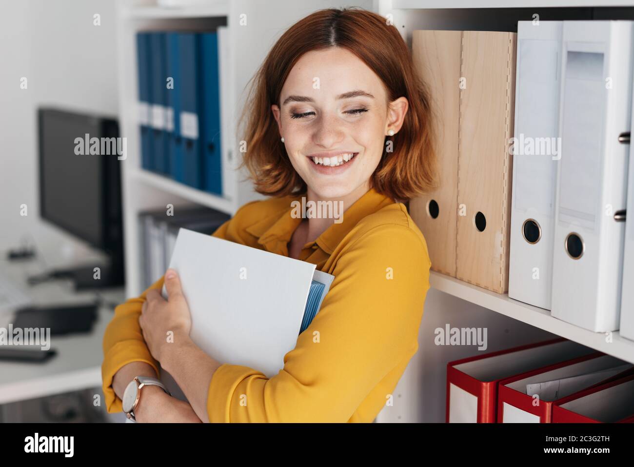 Young businesswoman with a wide smile standing clutching a large binder to her chest in the office with downcast eyes Stock Photo