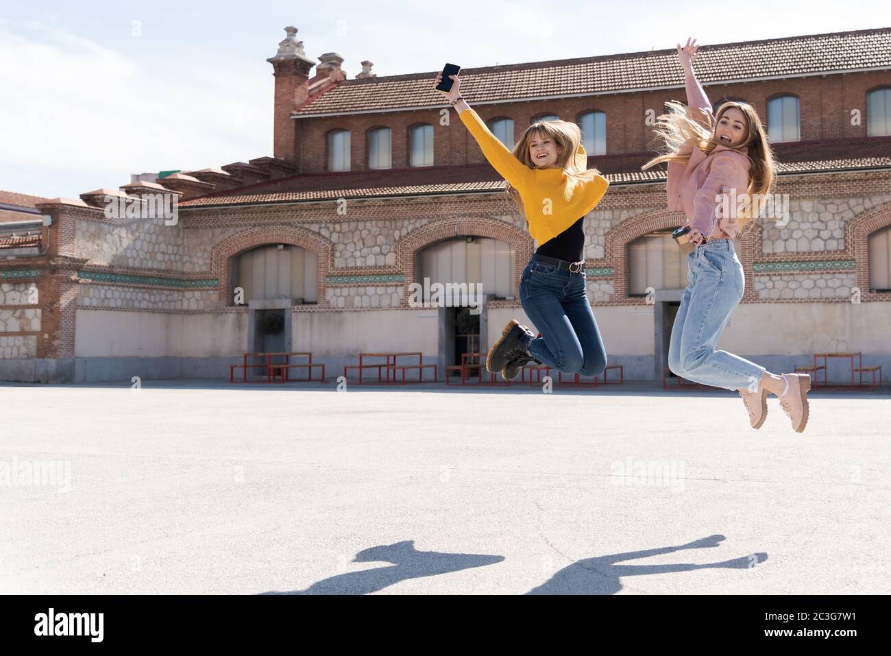 Two attractive young Caucasian girls jump up and down smiling in a street square. Bright image with natural light and space for text Stock Photo