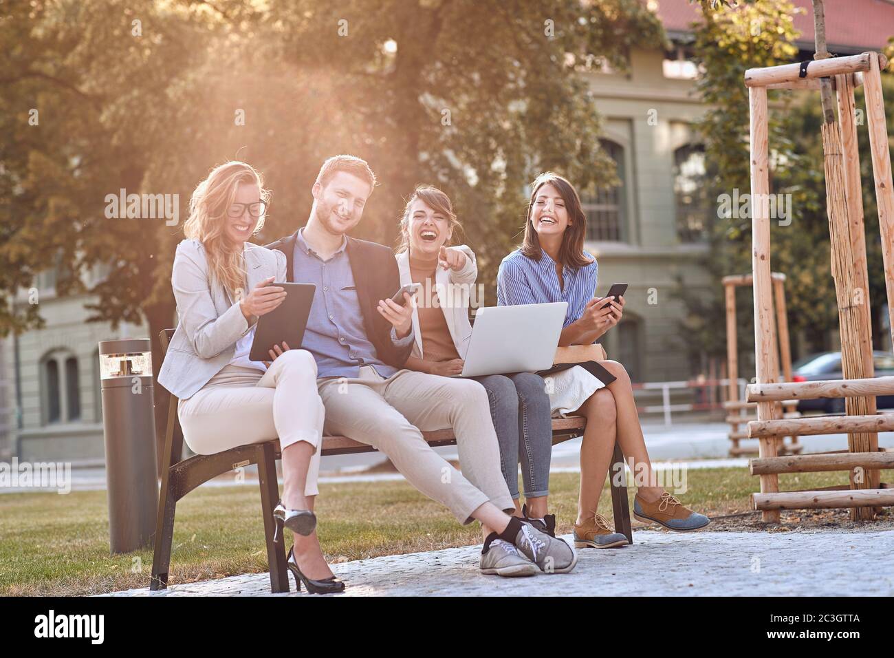 young caucasian businesspeople laughing at bench in a park with their cell phones, laptop, tablet. Looking at camera, eye contact. Break, pause from w Stock Photo