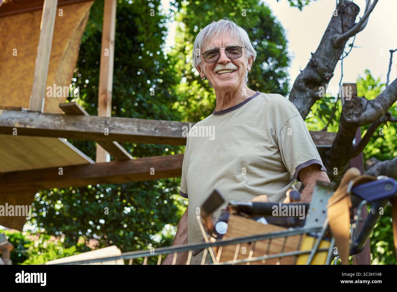 Retired happy senior Man in his 70s building a tree house at his back yard during warm summer day. Big smile showing nice teeth, gray hair. Stock Photo