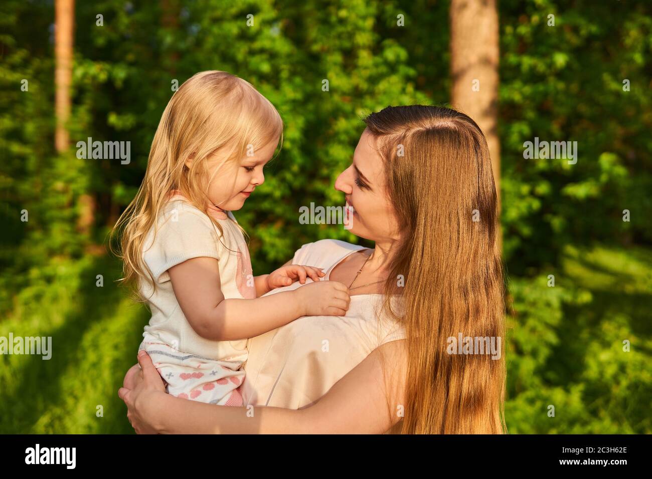 smiling beautiful young woman affectionately looks at her little daughter, who is holding in her arms, on a natural background Stock Photo