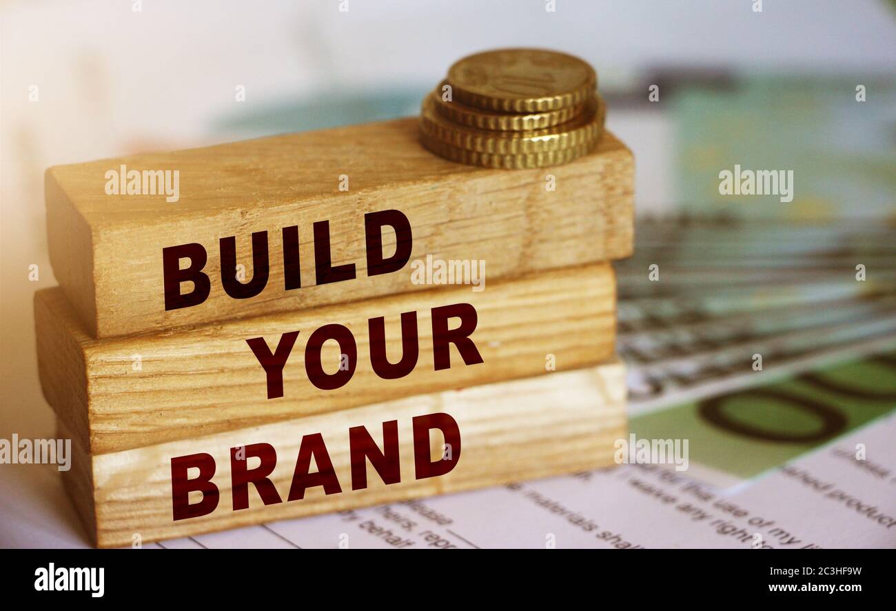 Wooden blocks form the text 'build your brend' on 100 Euro currency bills and coins background. Branding and marketing business concept Stock Photo