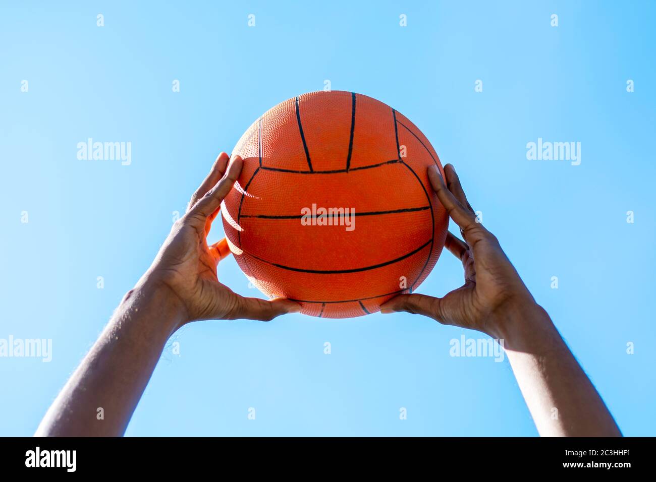 Close up shot of two dark-skinned hands holding up a basketball outdoors against blue clear sky, low angle. Stock Photo