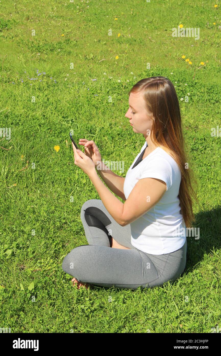 A young beautiful girl of European appearance in a white T-shirt with long blond hair sits on green grass, on the lawn with a mobile phone in her hands, flips through social networks, works in the phone. Stock Photo