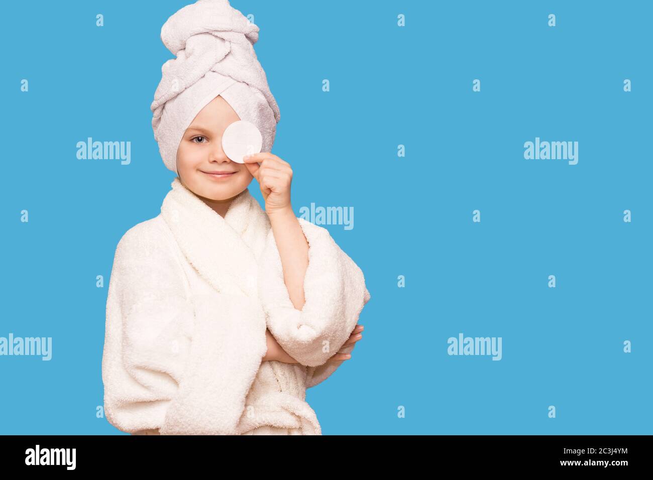 Smiling beautiful kid girl with towel on head hold cotton pad disk cleansing face skin with cleanser isolated on blue background. Happy girl remove ma Stock Photo