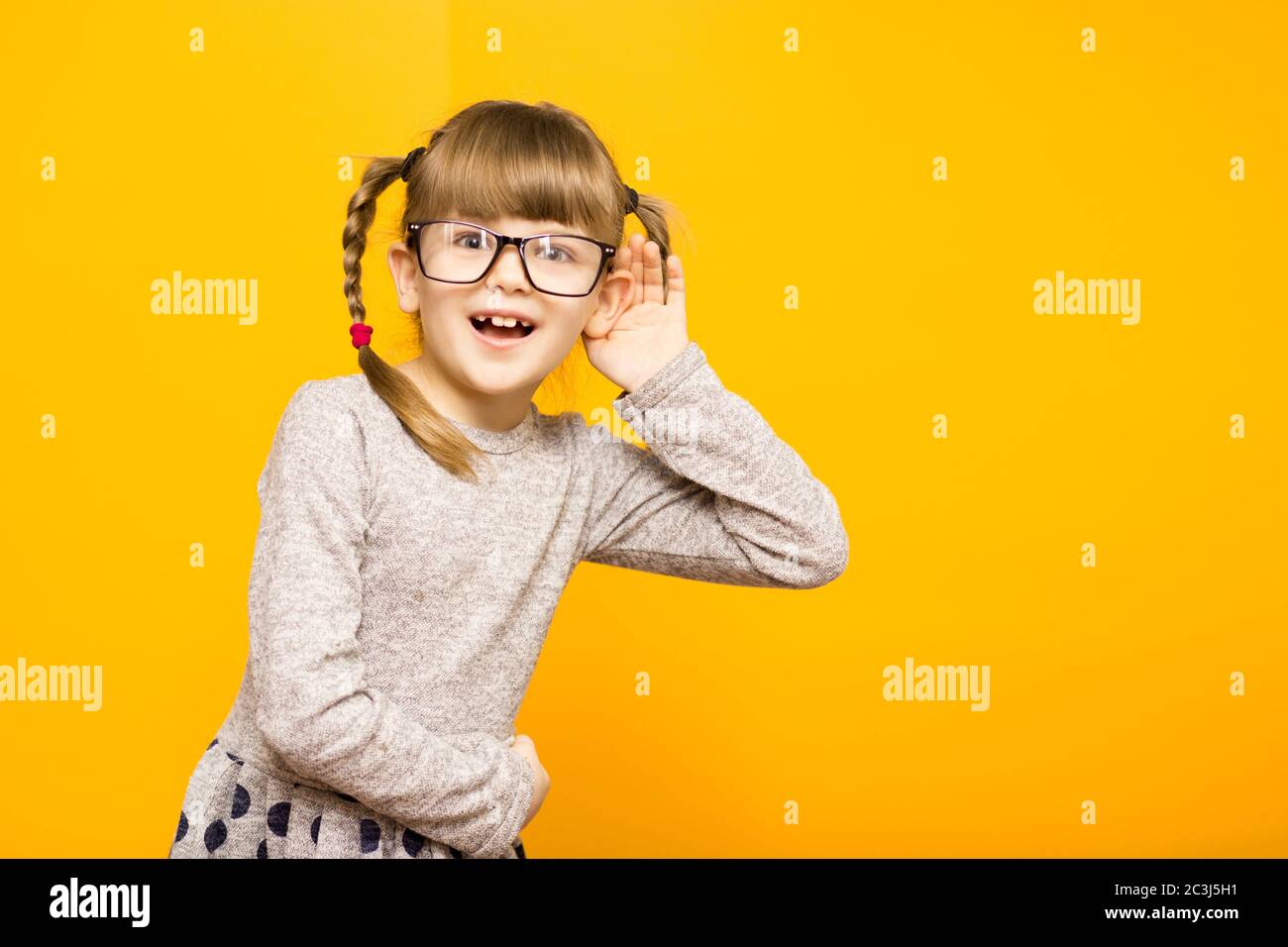 Little child girl journalist in glasses with surprised euphoria face and funny pigtails listening to something holding his hand to ear isolated on yel Stock Photo