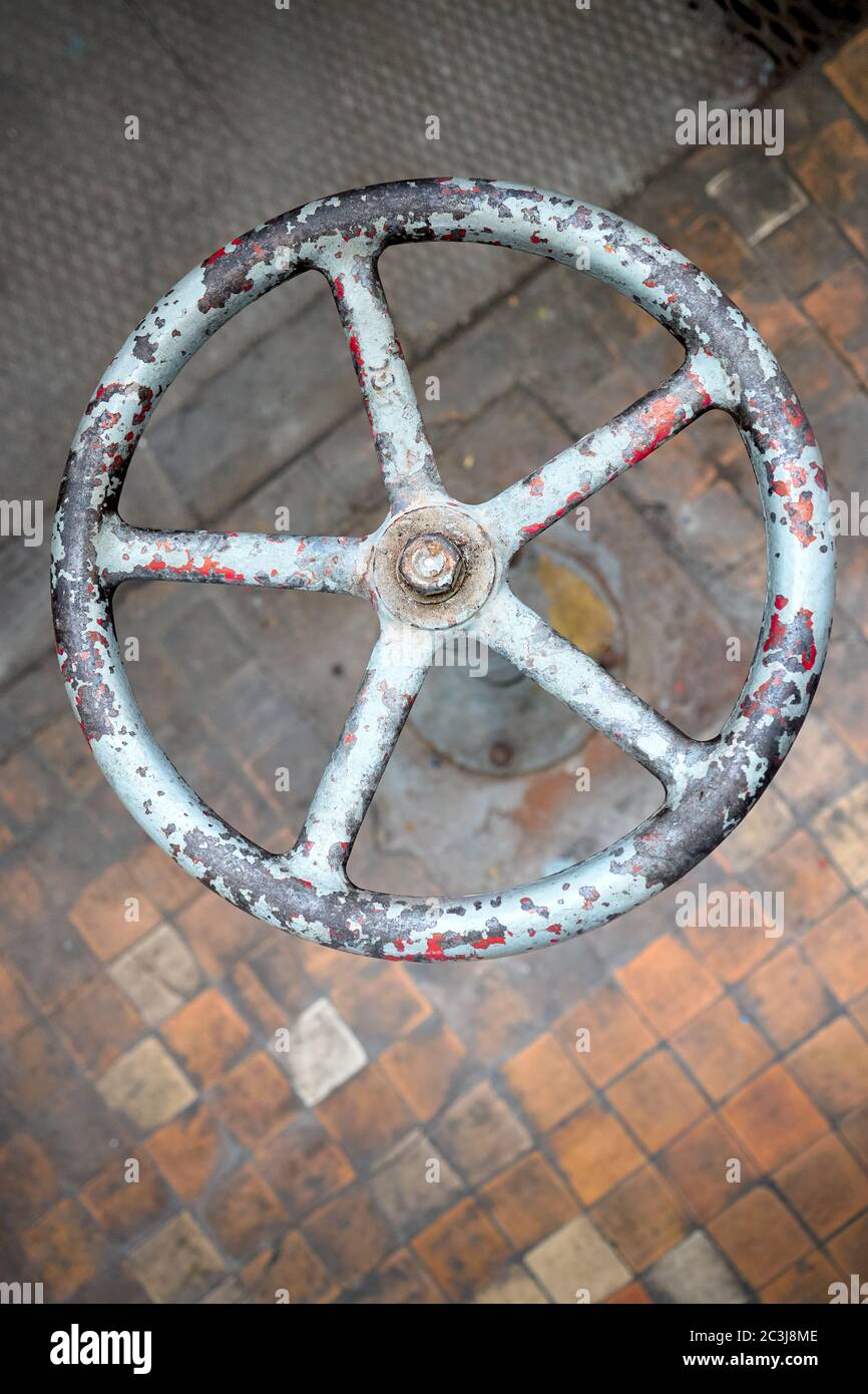 Old rusty valve wheel painted blue red with peeling selective focus with extension device over out of focus background tiled floor and perforated Stock Photo