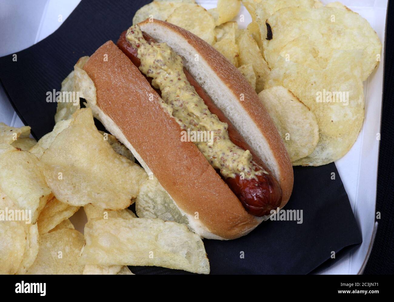 Hotdog covered in mustard inside a bun with potato chips on a black napkin Stock Photo