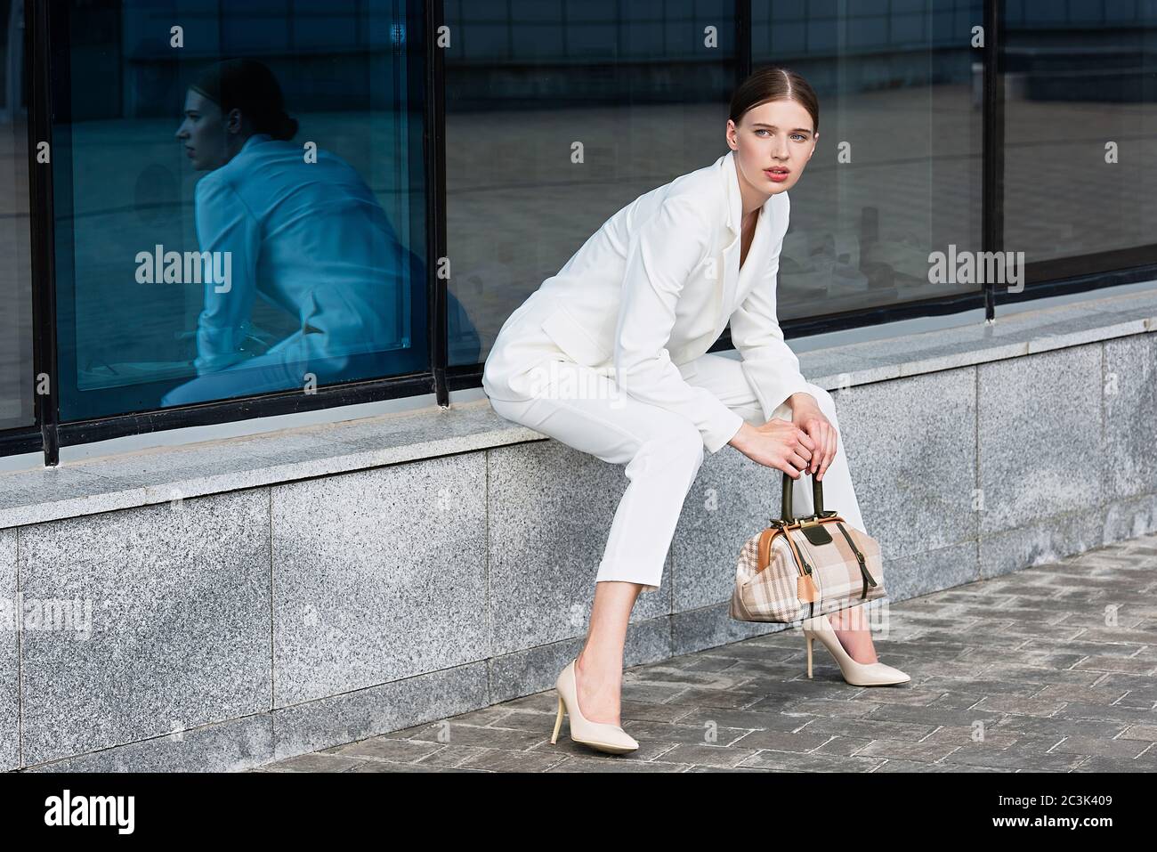 lifestyle girl in city, reflected in glass, girl holds  bag in her hands, white pantsuit and high heel shoes, Stock Photo