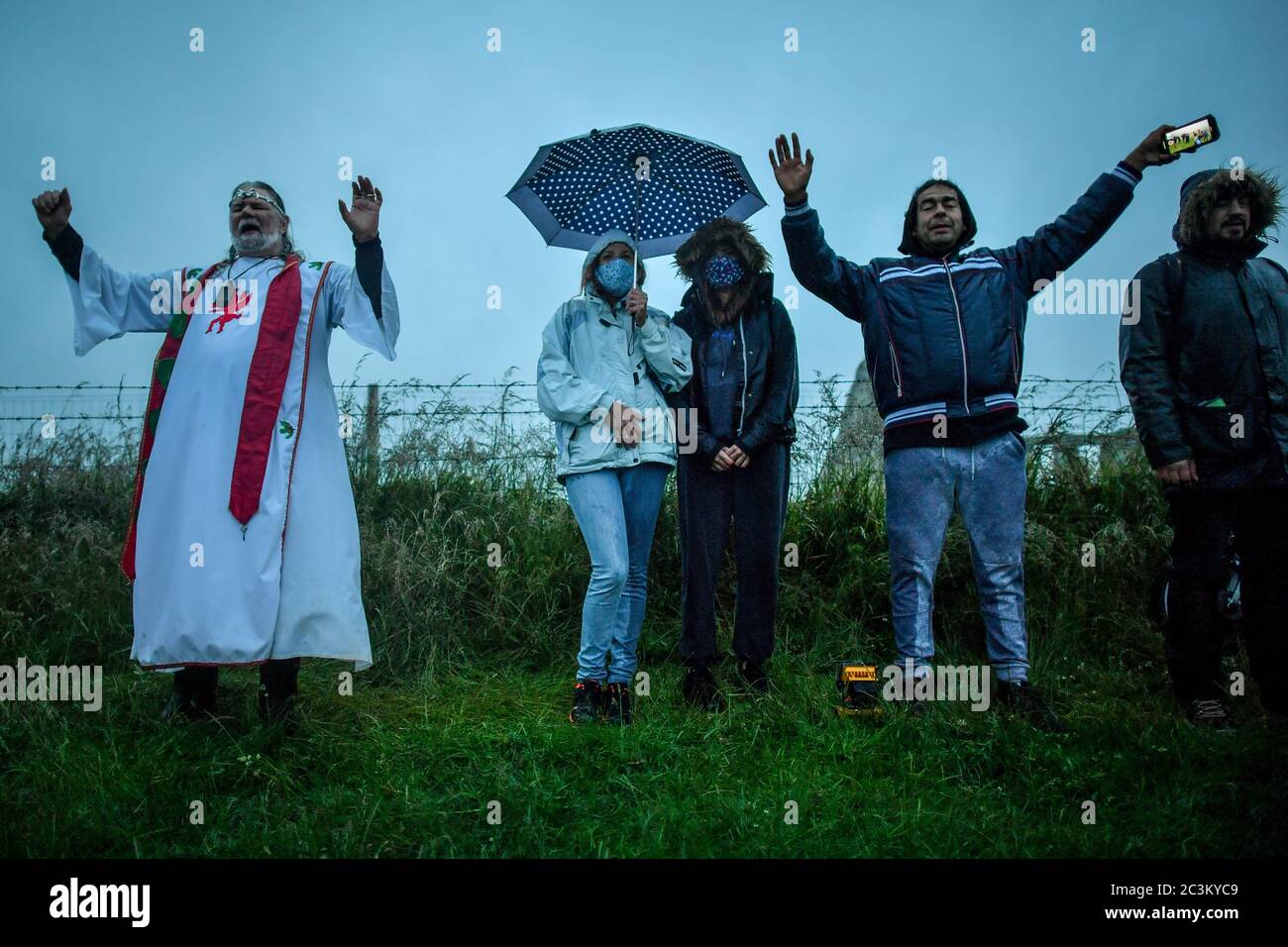 Arthur Uther Pendragon, left, and visitors turn to face the direction of the rising sun next to a closed Stonehenge as people gather to celebrate the dawn of the longest day in the UK. Stock Photo