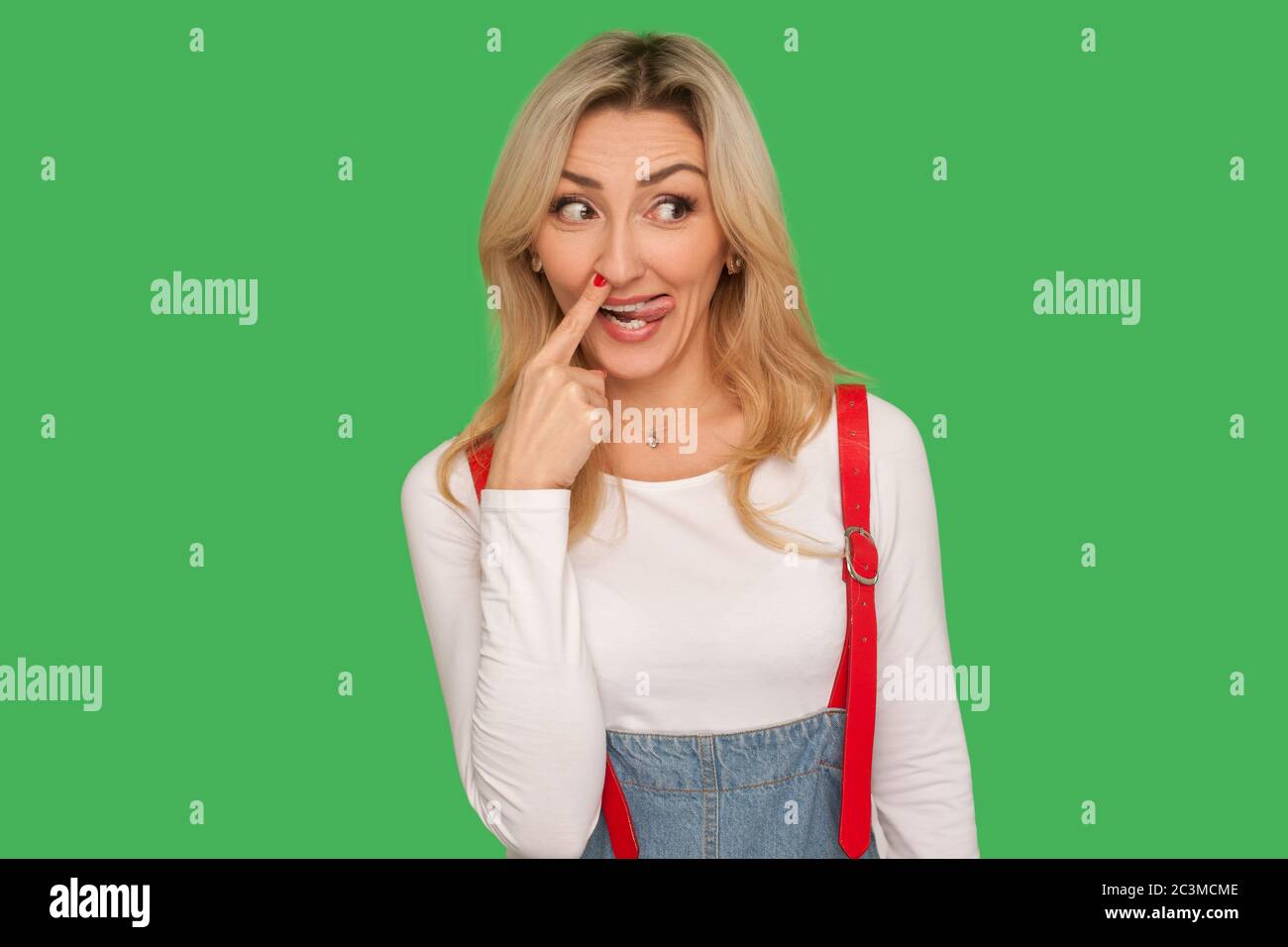 Portrait of crazy funny adult woman in denim overalls picking out boogers, drilling nose and smiling with comical humorous expression, fooling around. Stock Photo