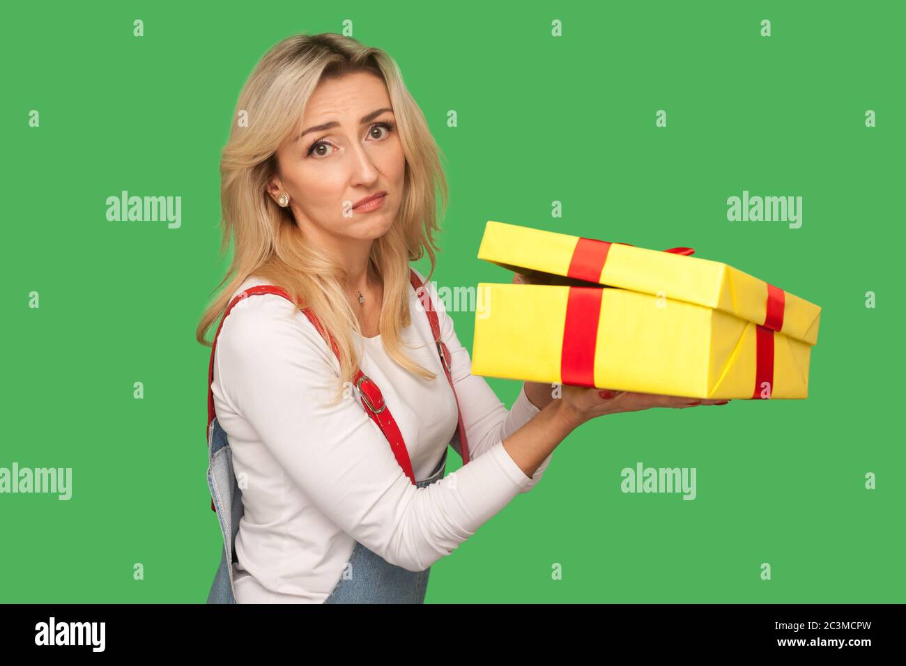 Portrait of upset displeased adult woman in denim overalls holding unwrapped gift, unpacking present and looking with disappointed sad expression. ind Stock Photo
