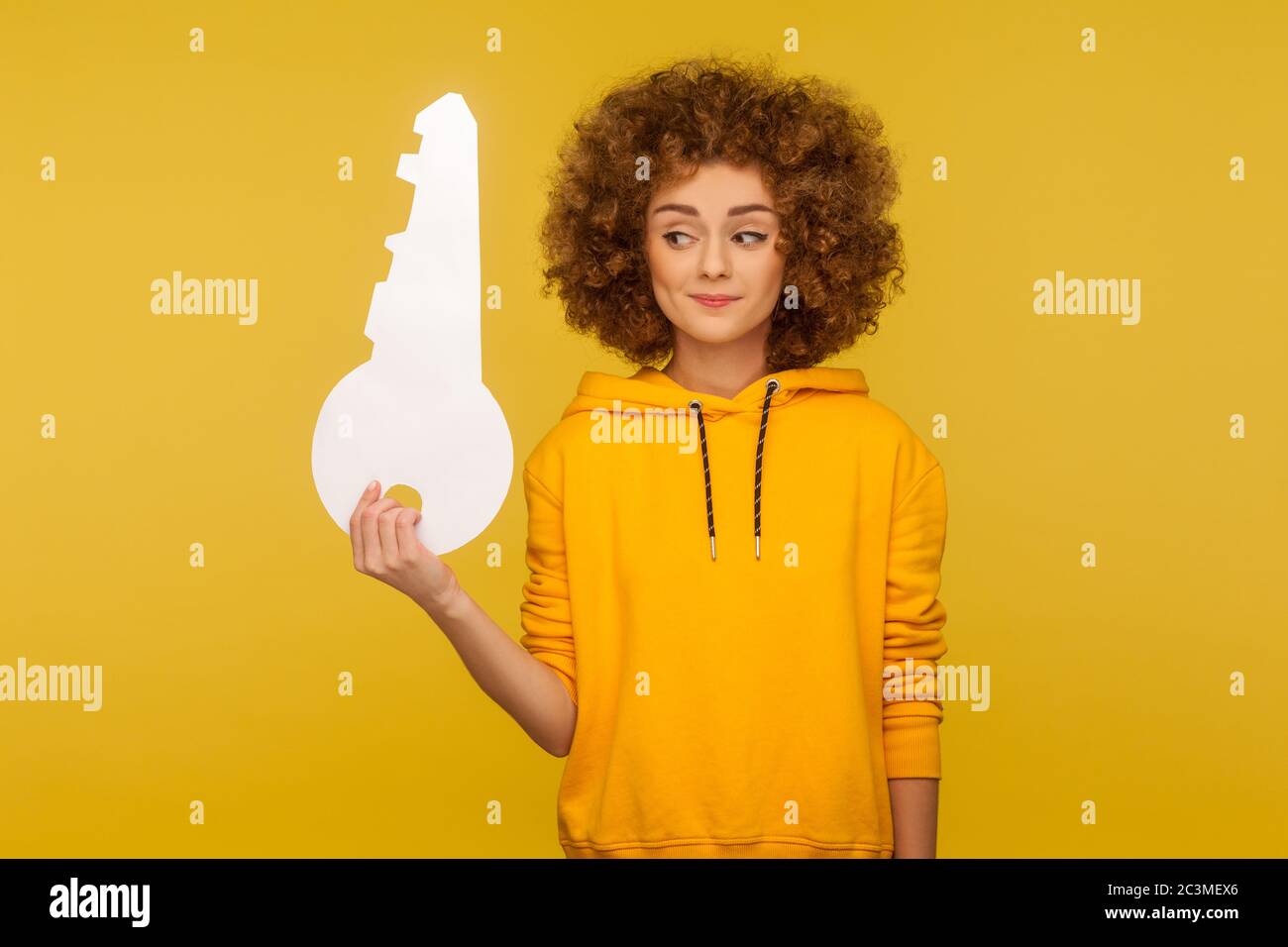 Portrait of funny curly-haired young woman in urban style hoodie looking at big paper key with interested curious expression, dreaming of own home. in Stock Photo