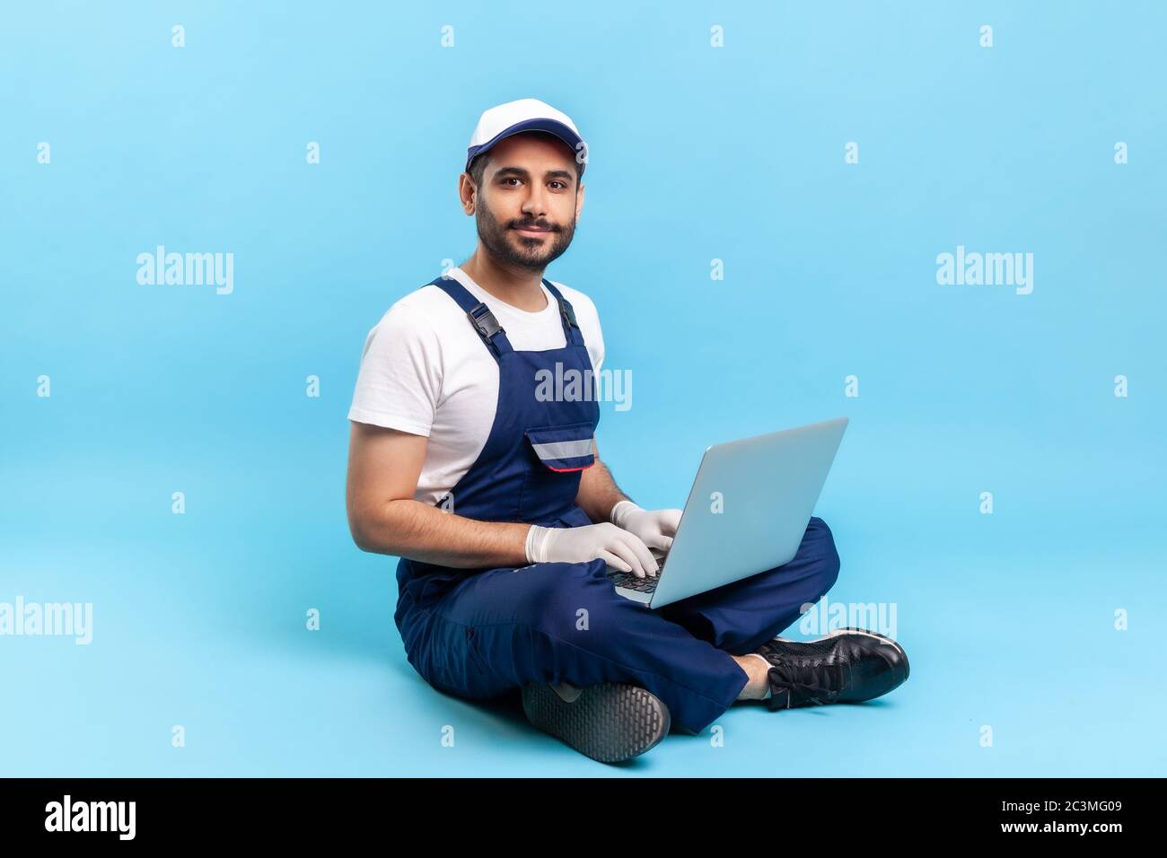 Repair service, online support. Portrait of happy professional handyman in blue workwear and protective gloves sitting cross-legged with laptop and sm Stock Photo