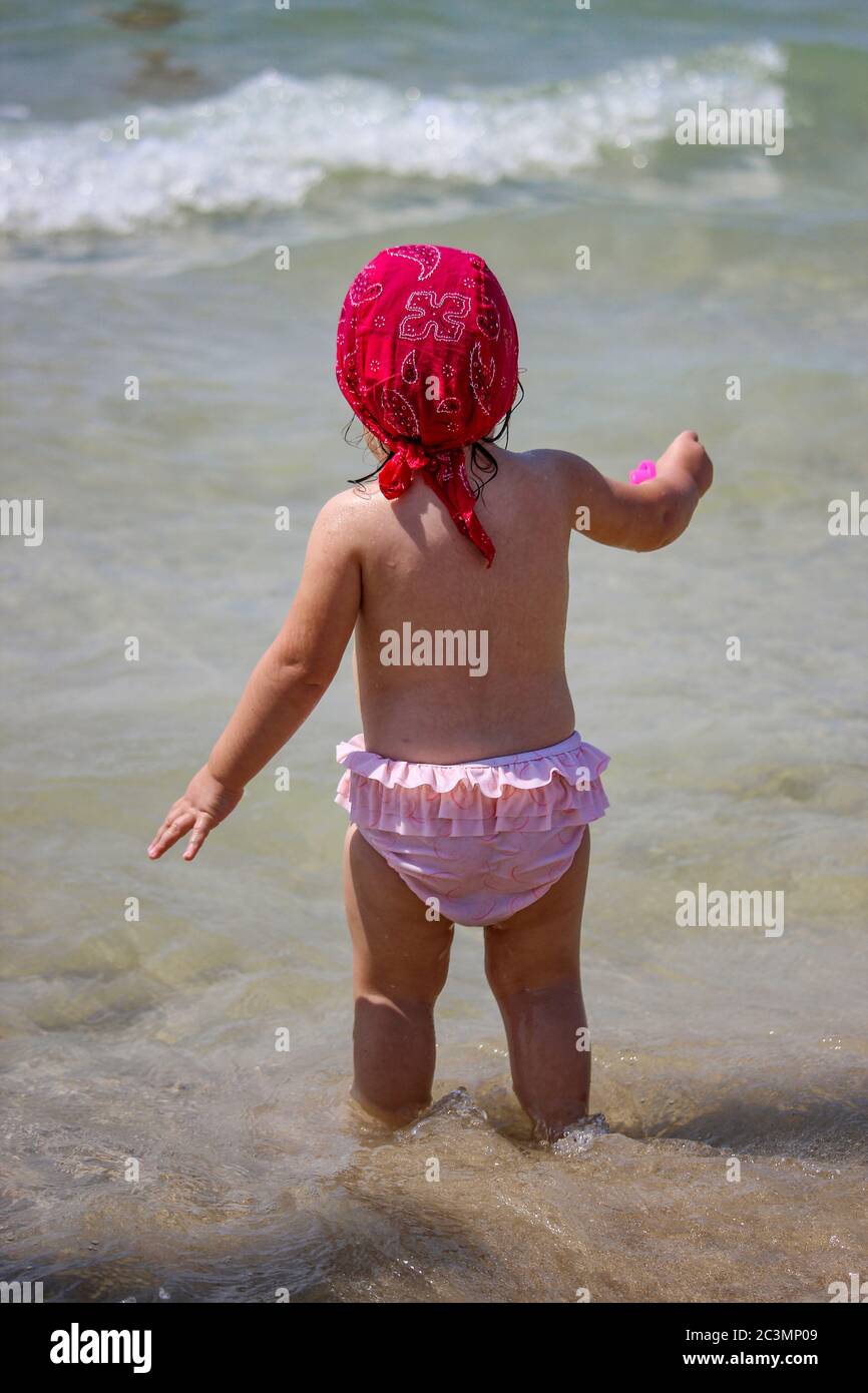 A cute and sweet little girl plays with water and sand on the seashore.  Child with pink swimsuit and red bandana, plays with a plastic scoop. Sea  foam Stock Photo - Alamy