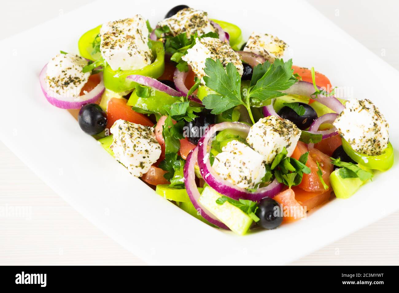 Greek salad with fresh tomato, cucumber, red onion, basil, feta cheese, black olives, Italian herbs and olive oil in white dish on white wooden table. Stock Photo