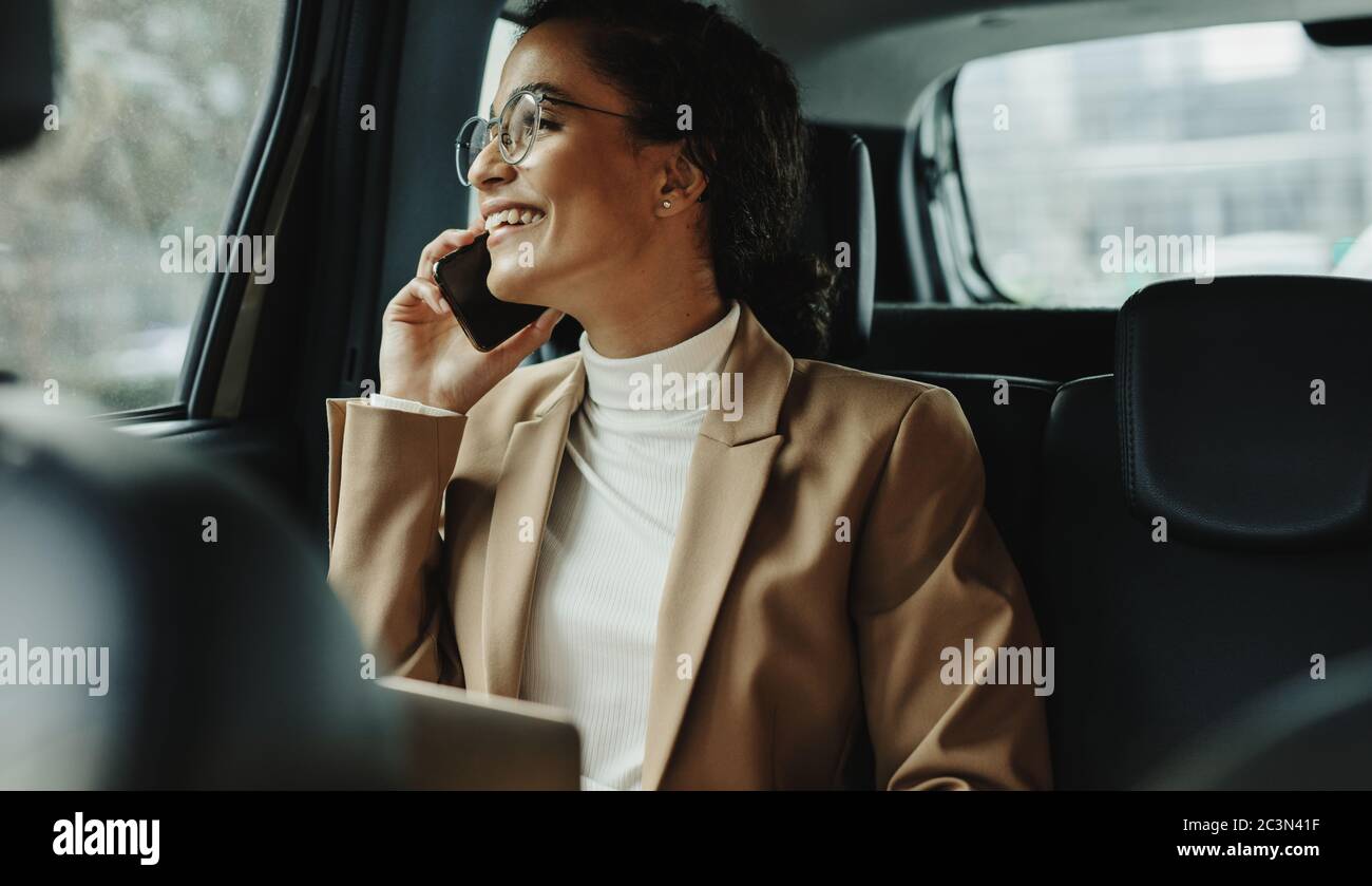 Businesswoman sitting on backseat of her car and talking on mobile phone. Businesswoman using phone while traveling by a car. Stock Photo