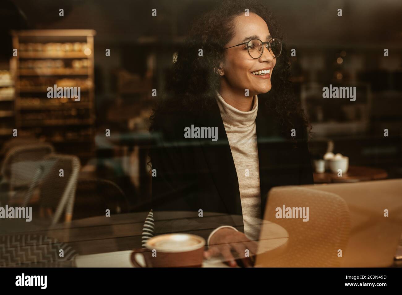 Businesswoman at a cafe with laptop looking outside the window and smiling. Female sitting at coffee shop with a her laptop. Stock Photo