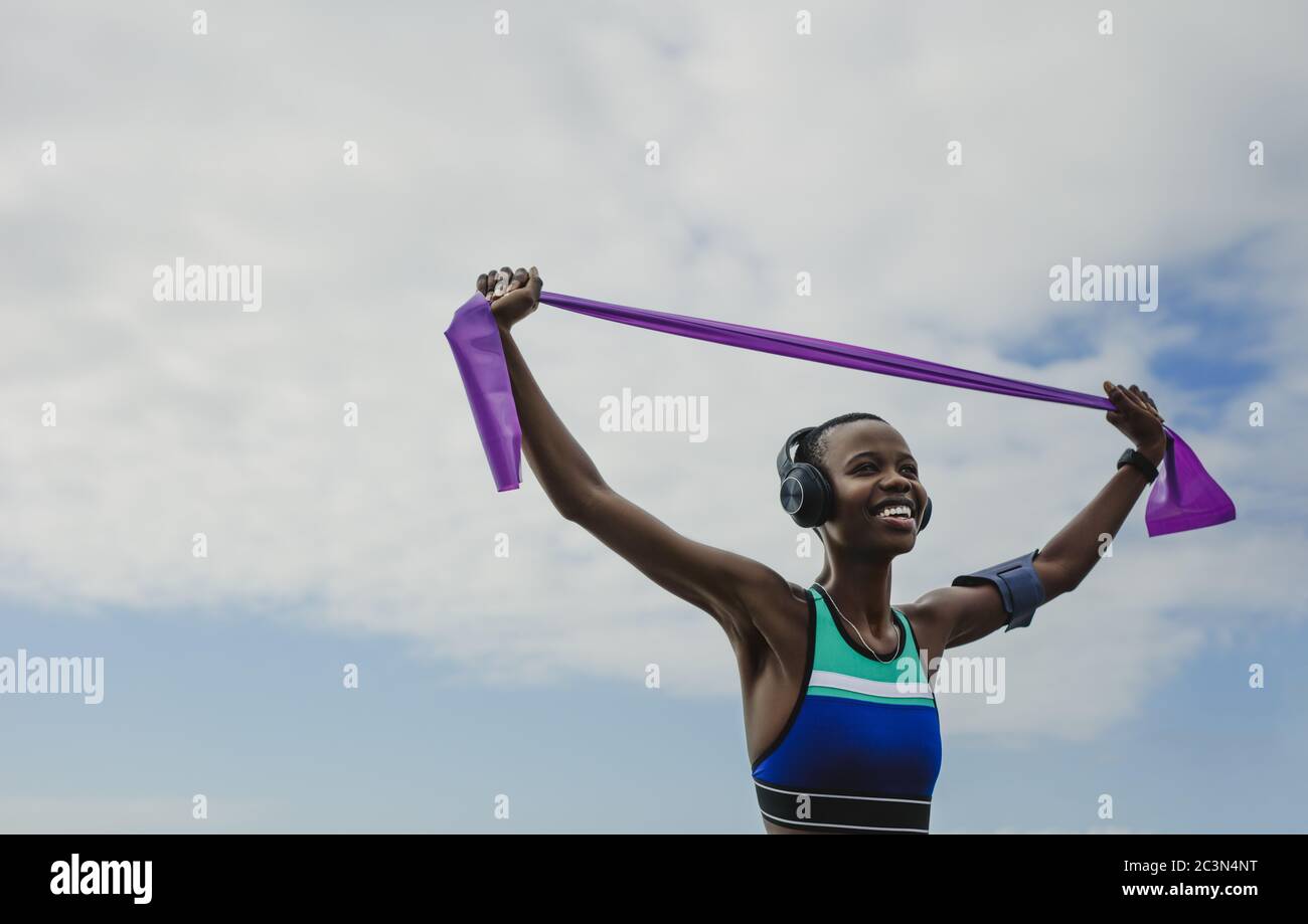 Happy woman in sportswear exercises with resistance band. Fitness female wearing headphones doing stretching workout against sky. Stock Photo