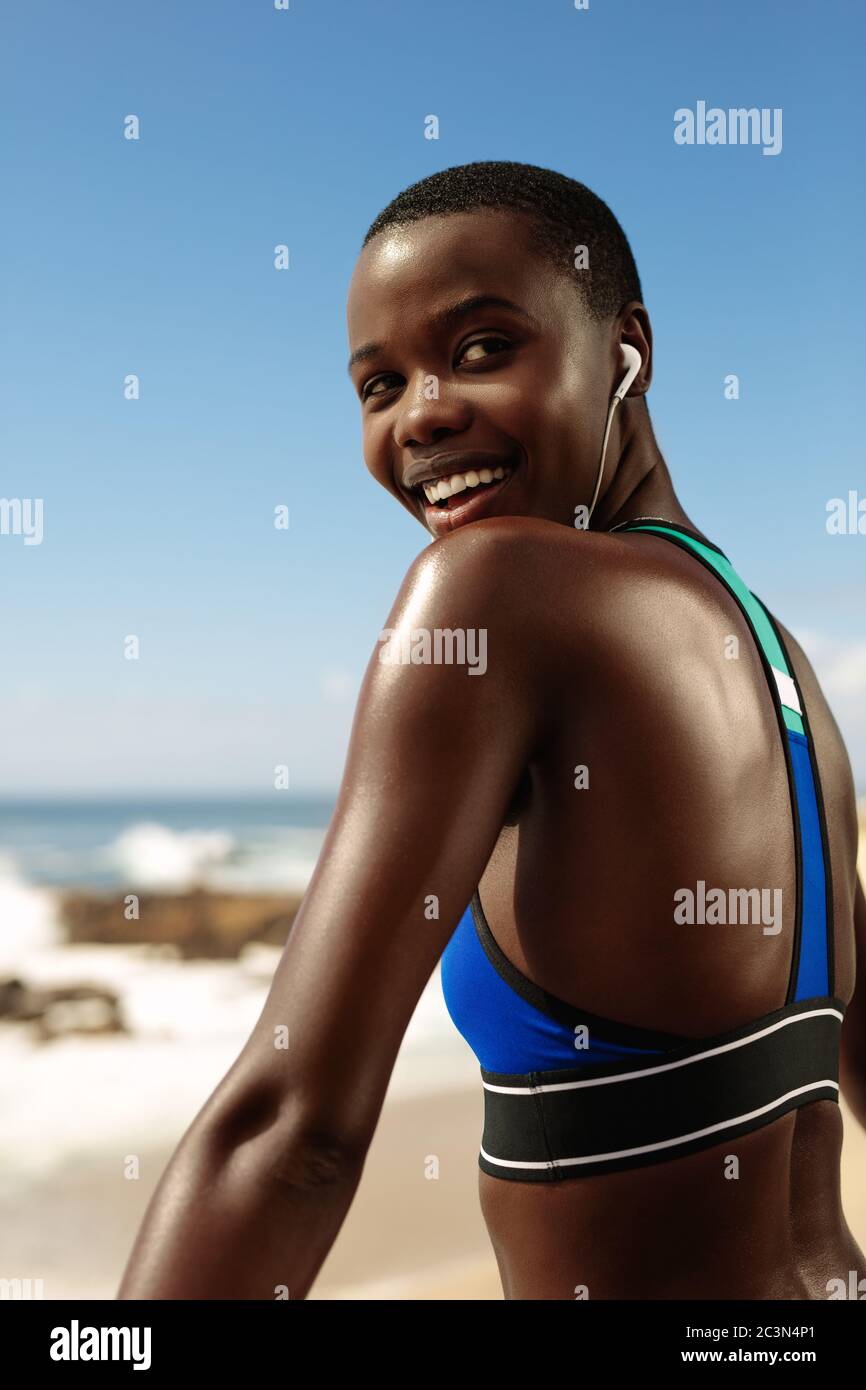African woman in sportswear taking a break after exercising outdoors. Fitness female wearing listening to music on earphones and smiling. Stock Photo