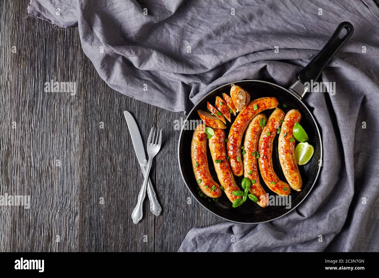 Original pork sausage links or patties fried on a skillet with fresh basil and spring onion served on a dark wooden background with cutlery, lime wedg Stock Photo