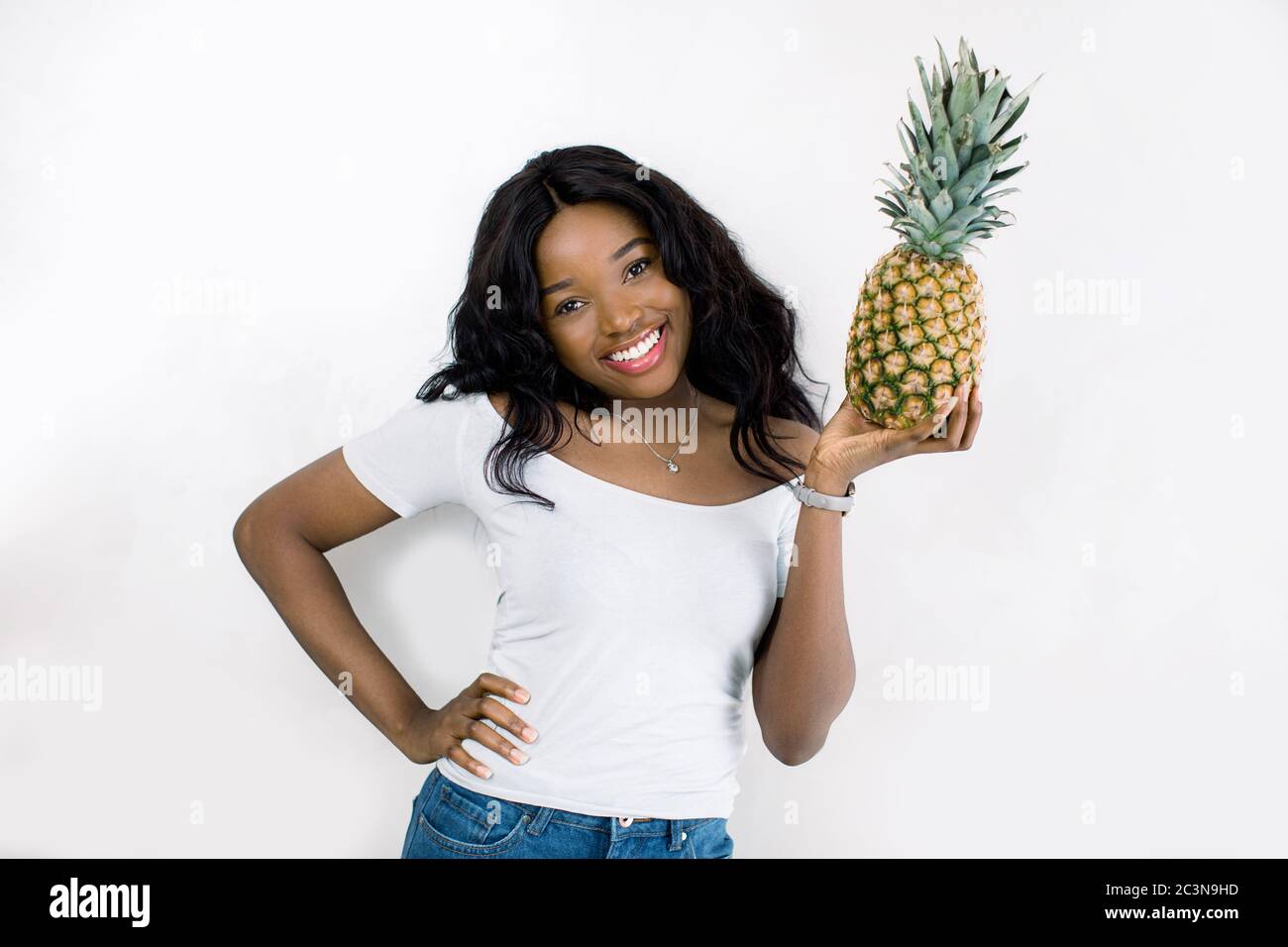 Gorgeous charming lovely young african girl with curly hair, in a white  t-shirt and jeans, holding a pineapple in hands, with a smile looks at  camera Stock Photo - Alamy