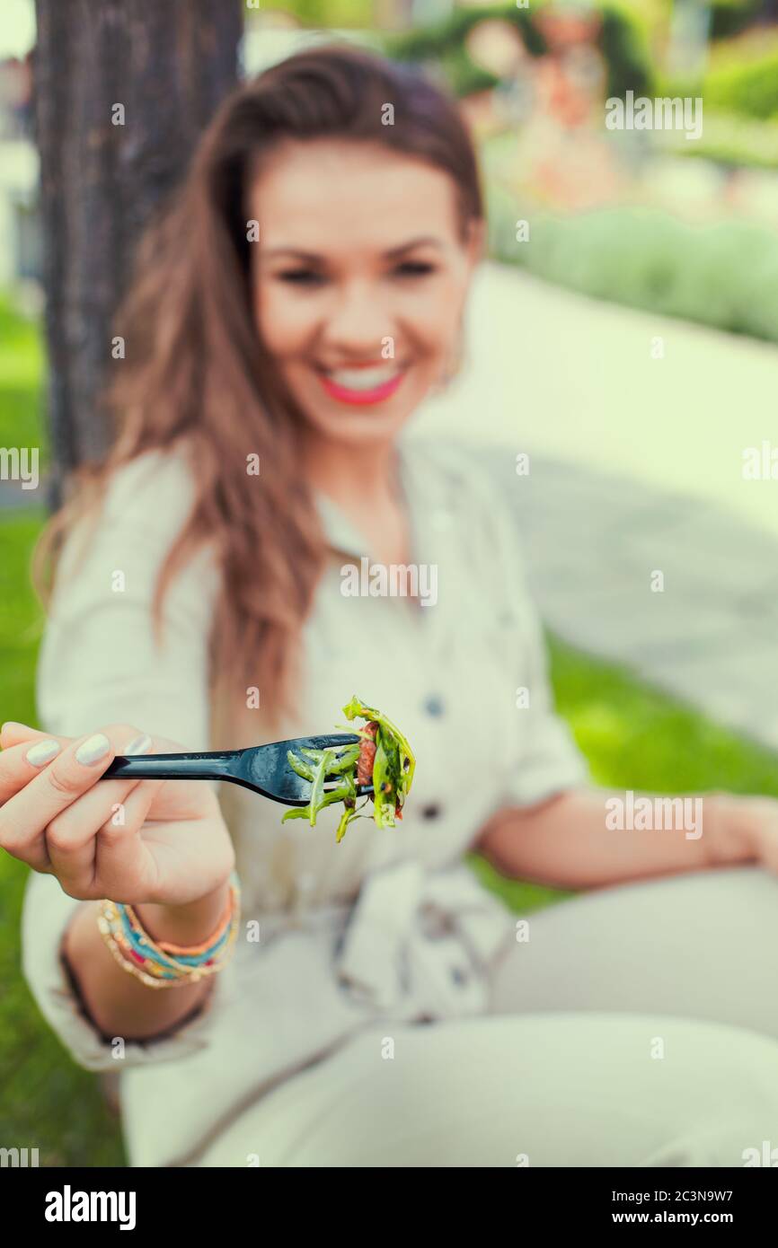 Happy young latina woman holding salad with toothy smile in park, depth of field Stock Photo