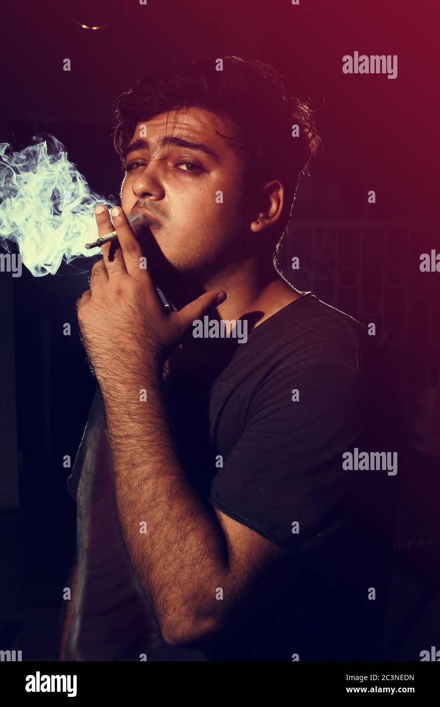 Young man smoking cigarette on dark background looking into camera wearing  t shirt Stock Photo